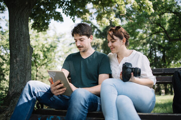 Canvas Print - Smiling couple looking at tablet while sitting on bench outdoor. Girl with DSLR camera sitting next to man who is holding tablet while they are sitting on bench in park.