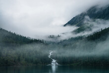 Highland Creek Flows Through Forest And Flows Into Mountain Lake. Ghostly Foggy Landscape With Alpine Lake And Dark Forest Among Low Clouds. Atmospheric Scenery With Coniferous Trees In Dense Fog.