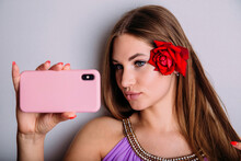 Attractive Young Girl Takes A Selfie On Her Smartphone, Pouting Her Lips And Straightening Her Hair With A Red Rose Flower.
