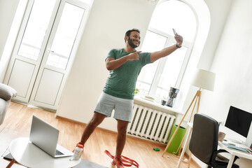 Wall Mural - Proud. Full length shot of excited bearded active man holding smartphone, pointing while taking a selfie during morning workout at home. Fitness, motivation concept