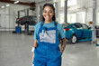 Perfect service. Portrait of young african american woman, professional female mechanic in uniform smiling at camera, standing in auto repair shop. Car service, repair, maintenance and people concept