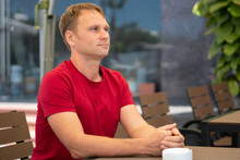 Half Side Portrait Restrained Smile Handsome Young Blond Man In Casual Red T-shirt Sit In Modern Cafe, Happy Look Away, Waiting Someone Or Met Already. Human Relationship, Good Future Changes Concept