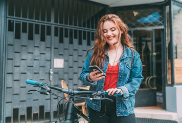 Wall Mural - Portrait of Red curled long hair caucasian teen girl on the city street walking with bicycle using the smartphone with earphones. Natural people beauty urban life concept image.