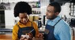 © VAKSMANV - African American handsome young male bartender in apron holding tablet device and female barrista writing list of orders or shopping at bar kitchen. Waiter and waitress making up note of menu for cafe