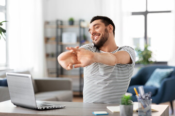 technology, remote job and business concept - happy smiling man with laptop computer stretching at home office