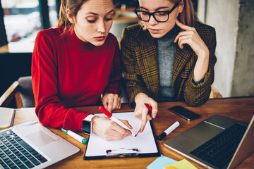 Cropped image of two skilled female designers collaborating on design sketch.Serious tutor pointing on blueprint helping student to drawing line during private lesson sitting at desktop with laptops