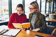 © BullRun - Two successful students reading textbook and studying together during exam preparation sitting in coworking.Serious female tutor with positive hipster girl conducting private lesson with literature