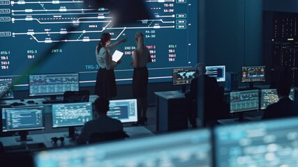 Poster - Two Software Engineers Talking Next to a Big Screen in a Modern Monitoring Office with Live Analysis Feed with Charts. Monitoring Room Big Data Scientists and Managers Sit in Front of Computers.