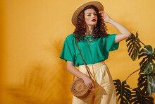 Summer Fashion Concept: Woman Wearing Trendy Green T-shirt, Yellow Bermudas Shorts, Straw Hat, With Round Shoulder Wicker Bag, Posing On Yellow Background. Copy, Empty Space For Text