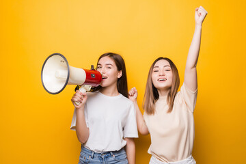 Two irritated young women girls friends scream in megaphone isolated on yellow background . People lifestyle concept.