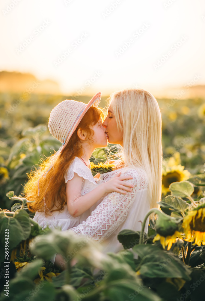 Happy mother kisses her little red-heired daughter in a field with yellow sunflowers on sunset. - obrazy, fototapety, plakaty 