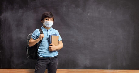 Wall Mural - Boy in school with a backpack and books standing in front of a school blackboard and wearing a face mask