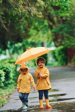 Two Kids With Umbrella In Yellow Waterproof Cloaks And Boots Playing Outdoors After The Rain Together