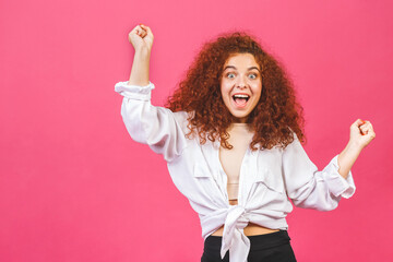 Photo of cheerful beautiful curly young woman standing isolated over pink wall background. Showing winner gesture.