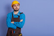 Shot of pensive dissatisfied construction worker dresses protective yellow helmet and brown apron, stands with arms folded, thinks over new building project.