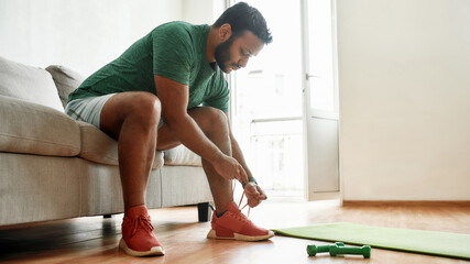 Almost ready. Young active man in sneakers tying shoelaces, preparing for morning workout with dumbbells on a yoga mat at home. Fitness, motivation concept