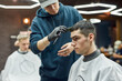© Svitlana - Side view of a professional male barber making new trendy haircut for a young handsome guy visiting barbershop or hair salon