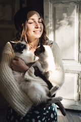 Wall Mural - Attractive girl embracing her Siamese cats. Indoor portrait of cute woman playing with her two pets.