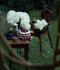 Brown fruit cake on the wooden narrow table in the garden. Silver tea pot and two black cups of tea are complementing the composition. There are white hydrangeas in the background.