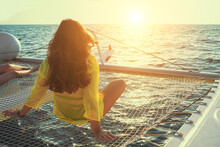 Woman Sits On The Deck Of A Sailing Catamaran At Sunset In The Sun. Rear View Of A Woman Enjoying The Azure Water On The Deck Of A Sailing Yacht. Selective Focus, Blur