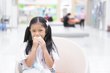 Wall Mural - Asian child hungry or kid girl eating bread or holding sandwich before learn at school for morning food or snack breakfast and hanging face mask on neck for coronavirus protect in department store