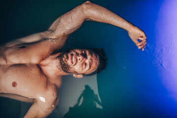 Wall Mural - Handsome beard man floating in tank filled with dense salt water used in meditation, therapy, and alternative medicine.