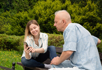 young blonde shows a man with gray hair something on the phone screen. they sit on a park bench