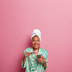 Sticker - Vertical shot of beautiful dark skinned woman eats delicious ice cream with spoon, concentrated above, dressed in domestic outfit, enjoys summer holidays at home, isolated over rosy wall, blank space