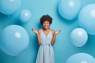 Wall Mural - Good looking curly haired woman spreads palms, smiles sincerely, enjoys summer party, wears blue dress, stands against festive air balloons, has happy mood, isolated. Feminity, style, fashion concept