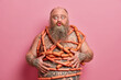 © wayhome.studio  - Funny fatso male keeps lips rounded, poses with naked body wrapped by sausages, has health problems connected with health and unhealthy nutrition, likes meat, stands indoor. Gluttony concept