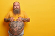 © WHstudio Leushin N - Photo of overweight pensive man keeps hands on big belly with tattoo, thinks and looks aside, has thick beard, poses against yellow background. Obese guy unable to realize how tummy could appear
