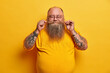 © WHstudio Leushin N - Indoor shot of pleased plump man twirls mustache, boasts of thick beard, stands with closed eyes, smiles pleasantly, has tattooed arms dressed in yellow clothes wears round small glasses poses indoor