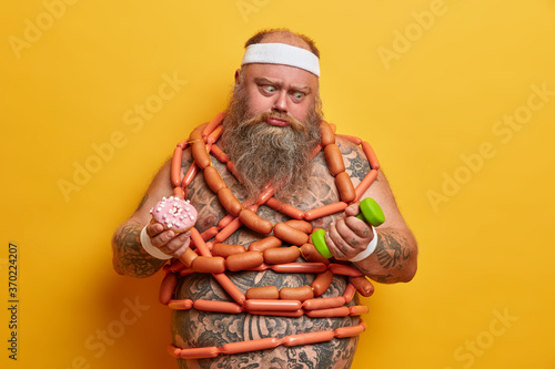 Cuadros en Lienzo Overweight man has temptation to eat junk food goes in for sport for loosing weight has intensive cardio workout in gym holds dumbbell, doughnut cannot stop eating fast food