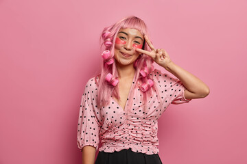 Wall Mural - Happy pink haired girl applies eye patches for reducing dark circles, makes peace sign, wears hair curlers to have perfect curls, has fun, isolated over rosy background. Monochrome. Skin treatment