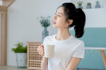 Wall Mural - Young asia woman with ponytail in white t-shirt holding a cup of coffee or tea relaxing at home in the morning.