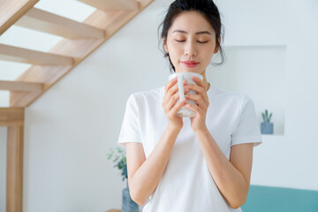 Wall Mural - Young asia woman with ponytail in white t-shirt holding a cup of coffee or tea relaxing at home in the morning.