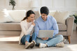 © fizkes - Smiling young family couple sitting on floor in living room, looking at laptop screen. Happy millennial woman and man planning holiday vacation honeymoon trip, shopping online or watching movie.