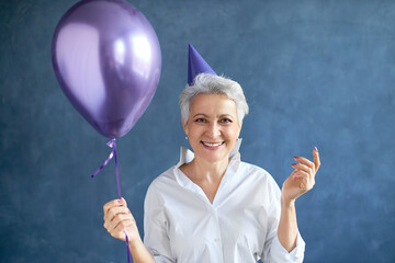 Isolated image of attractive successful mature businesswoman with short gray hair posing at blank studio wall with helium balloon, expressing positive emotions ar surprise party. People and lifestyle