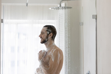 Smiling young man taking morning shower, standing in modern bathroom, applying natural moisturizing foamy gel or shampoo, holding fluffy bath puff, personal hygiene and skincare concept