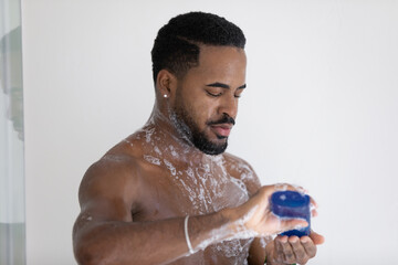 Close up handsome African American young man taking shower with moisturizing gel or shampoo, applying cosmetic product, holding plastic bottle, enjoying morning routine, skincare procedure