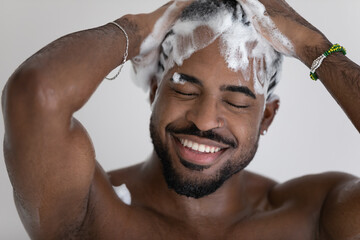 Close up smiling African American handsome young man washing cleaning hair with foamy anti-dandruff shampoo, taking shower, enjoying morning routine procedure, standing in bathroom