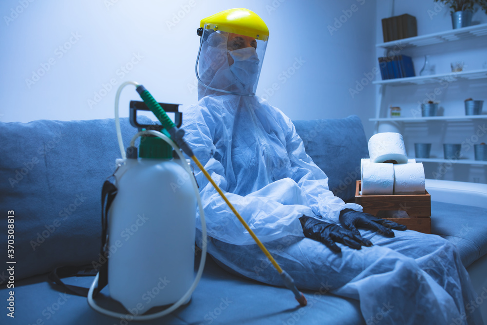 Person with toilet paper stock and protective antiviral mask, suit, helmet and chemical decontamination sprayer bottle in home isolation. - obrazy, fototapety, plakaty 