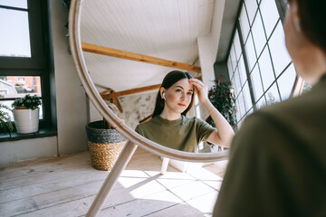 Wall Mural - Attractive young caucasian woman in headphones touching face and looking at mirror in living room at home in sunny day. Beauty and skin care concept.