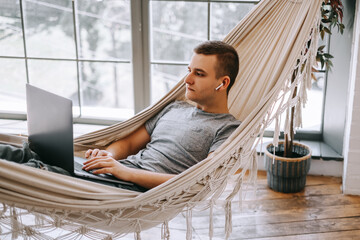 A young caucasian businessman in headphones works on a laptop, browses the internet while lying in a hammock at the home office. A person working on electronic devices.