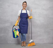© Friends Stock - Happy cleaning lady in uniform and yellow rubber gloves holding broom and plastic bucket with rags and different cleaning products while standing against grey wall