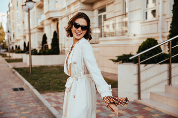 Wall Mural - Good-humoured girl in long coat smiling on the street. Well-dressed european woman spending time in city.