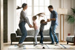 © fizkes - Full length positive energetic young parents holding hands of little kids siblings, jumping together in living room. Overjoyed family couple involved in dancing activity with small children at home.