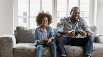 Wall Mural - Overjoyed young biracial father sit on sofa have fun play video games with cute little daughter, happy african American dad and small girl child engaged in funny activity together on weekend