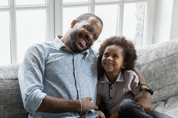 Wall Mural - Happy young biracial father sit relax on couch in living room cuddle hug with small ethnic son, overjoyed african American dad and biracial boy child enjoy family weekend at home, bonding concept