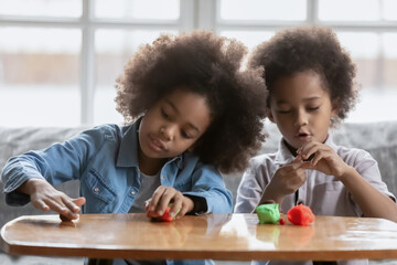 Little african american girl and boy kids have fun play with plasticine or modeling clay at home together, small children siblings engaged in creative activity with colorful playdough, hobby concept